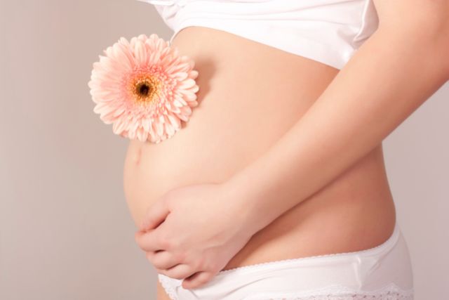 Une femme enceinte de 8 mois est de profil avec une main posée sur le bas de son ventre. Une grosse fleur rose est déposée au dessus de son nombril. La femme en question porte une brassière et une culotte de couleur blanche